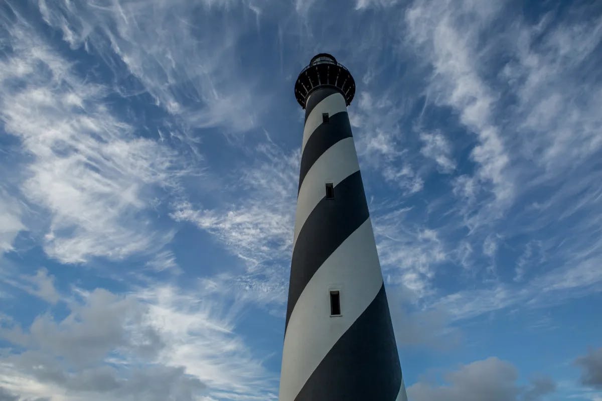 A black and white spiral striped lighthouse towering over with a cloudy blue sky in the background. 