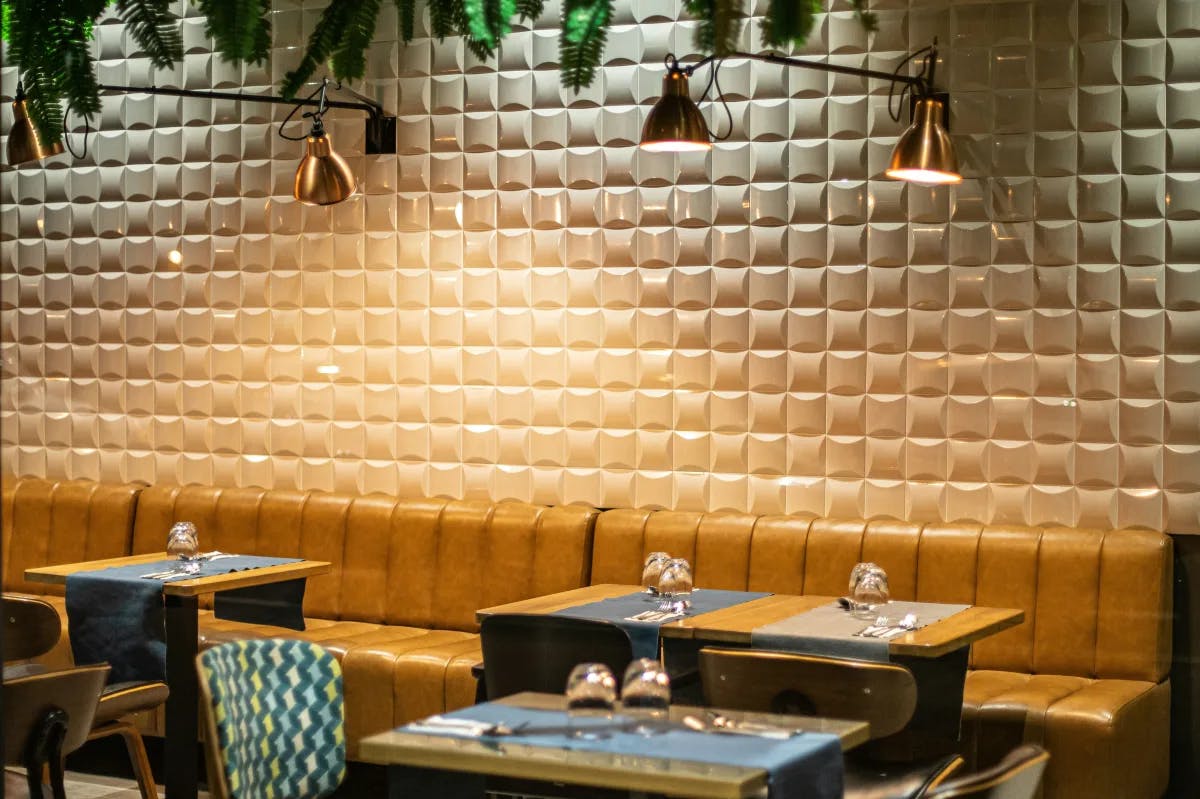 A restaurant with tiled walls, accent lighting, tan seating, wooden tables and patterned chairs. 