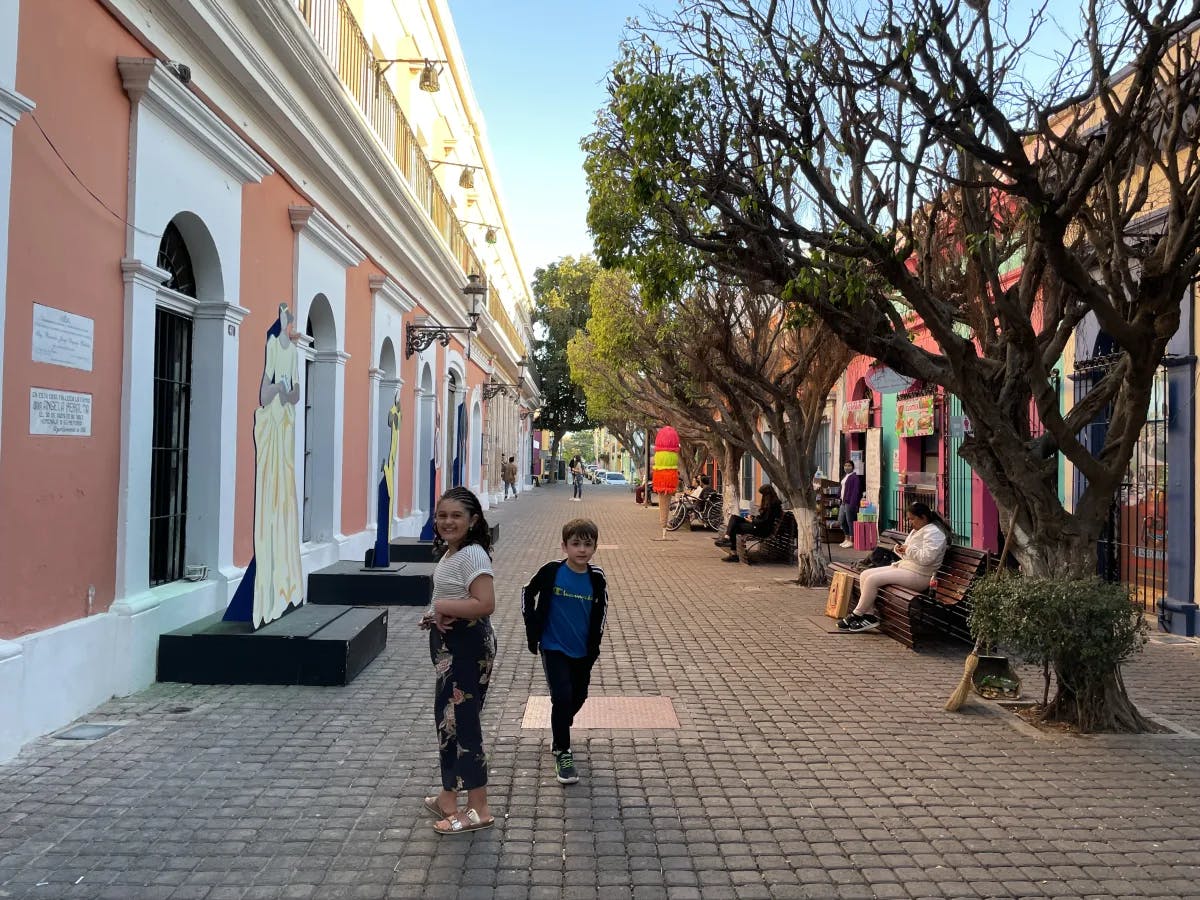 Two kids running down a street next to trees and arched buildings. 