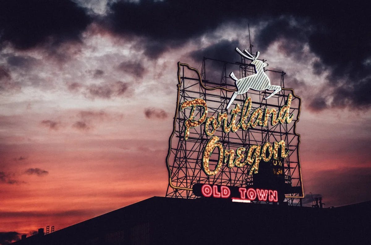 A view of a neon sign that reads Portland, Oregon with a white deer above it. There is a vibrant pink sunset and dark clouds hanging above in the background. 