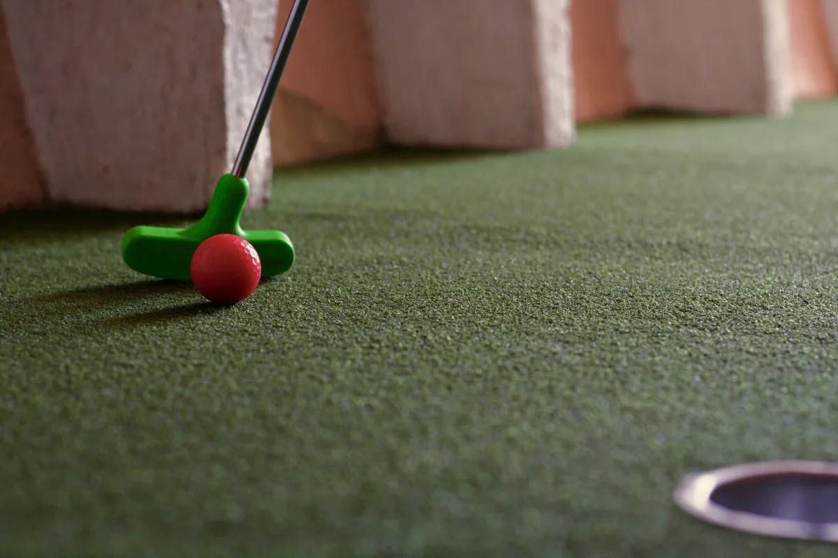 A close-up of a green mini golf putter and red ball on a course.