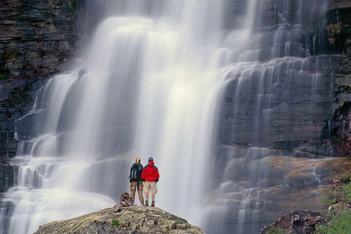 Two people standing on a rock in front of a large, cascading waterfall. One person is wearing a red jacket and khaki shorts while the other person is wearing a grey top, black backpack and khaki pants. 