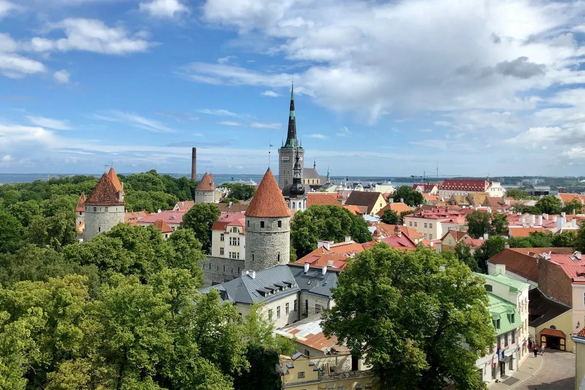 Tallinn Old Town is boasting medieval charm with cobblestone streets, historic buildings, and vibrant cafes in Estonia's capital city. Photo is an aerial view of the town with red-roofed buildings and a forest to the side.