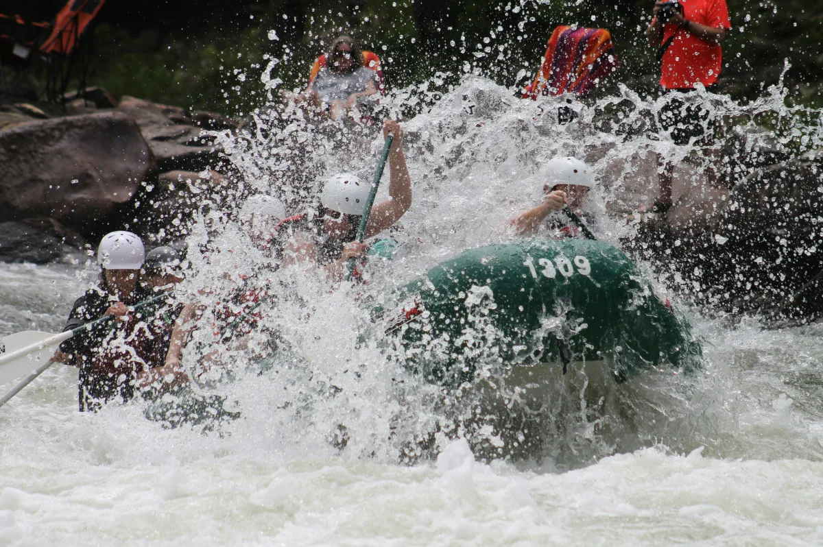A group of people whitewater rafting through rapids on a green raft.