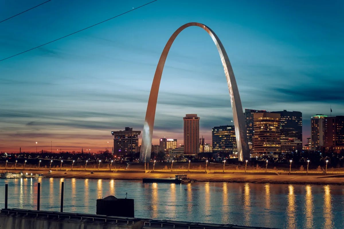 A view of the river and large silver arch in front of the city skyline at night. 