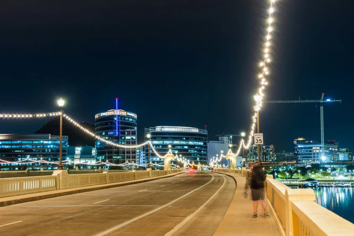 A view of a city bridge with string lights hanging down the sides, a person walking and a lit up skyline at nighttime. 