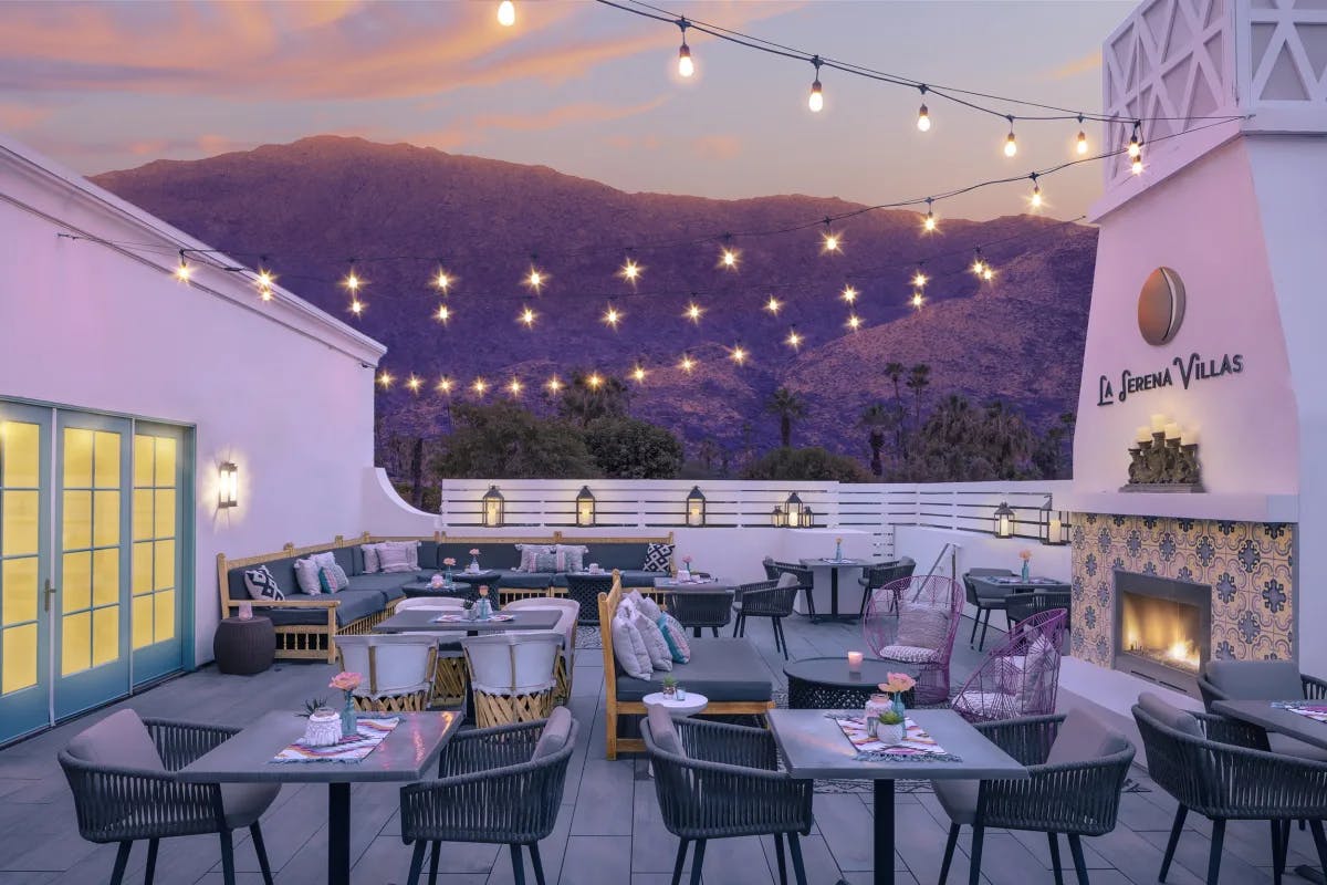 An image of a restaurant patio at sunset with string lights, a stone white fireplace and mountains in the background.