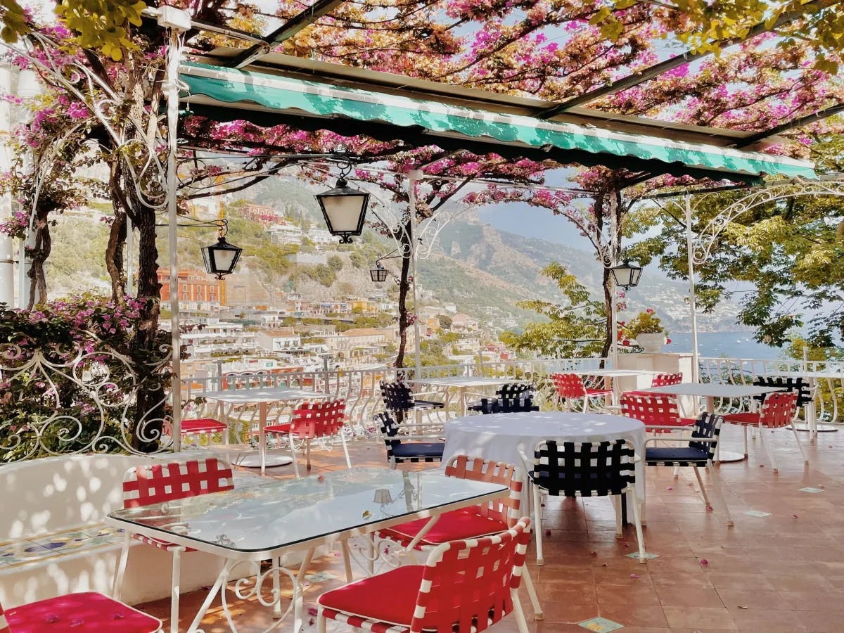 A picture of an outdoor terrace with red and black chairs, white tables, a pergola with flowers and vines draped over it, hanging lanterns and a view of the coastal town of Positano and the sea. 