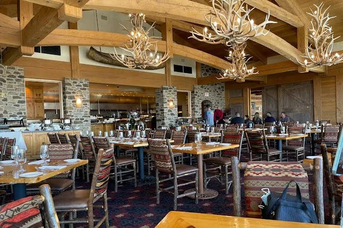 The interior of a restaurant with antler-style chandeliers, wood details, stone pillars and various dining tables. 