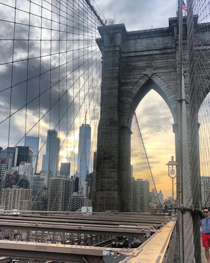 A view of the Brooklyn Bridge's beams and stone archways with the city skyline in the background and people walking in the forefront. 