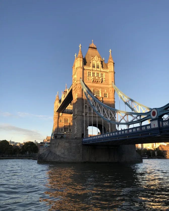 A view of a bridge in London with a large stone tower hanging over a reflective river. The sun is shining onto the tower. 