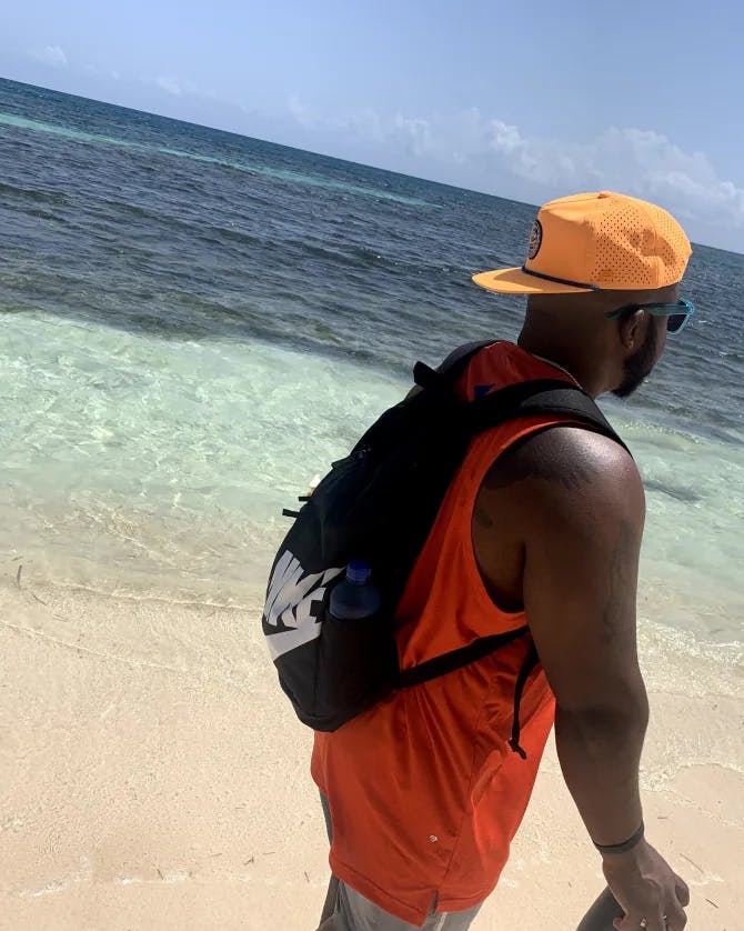 Picture of Harold wearing a tank top and orange hat walking on a beach with crystal clear water