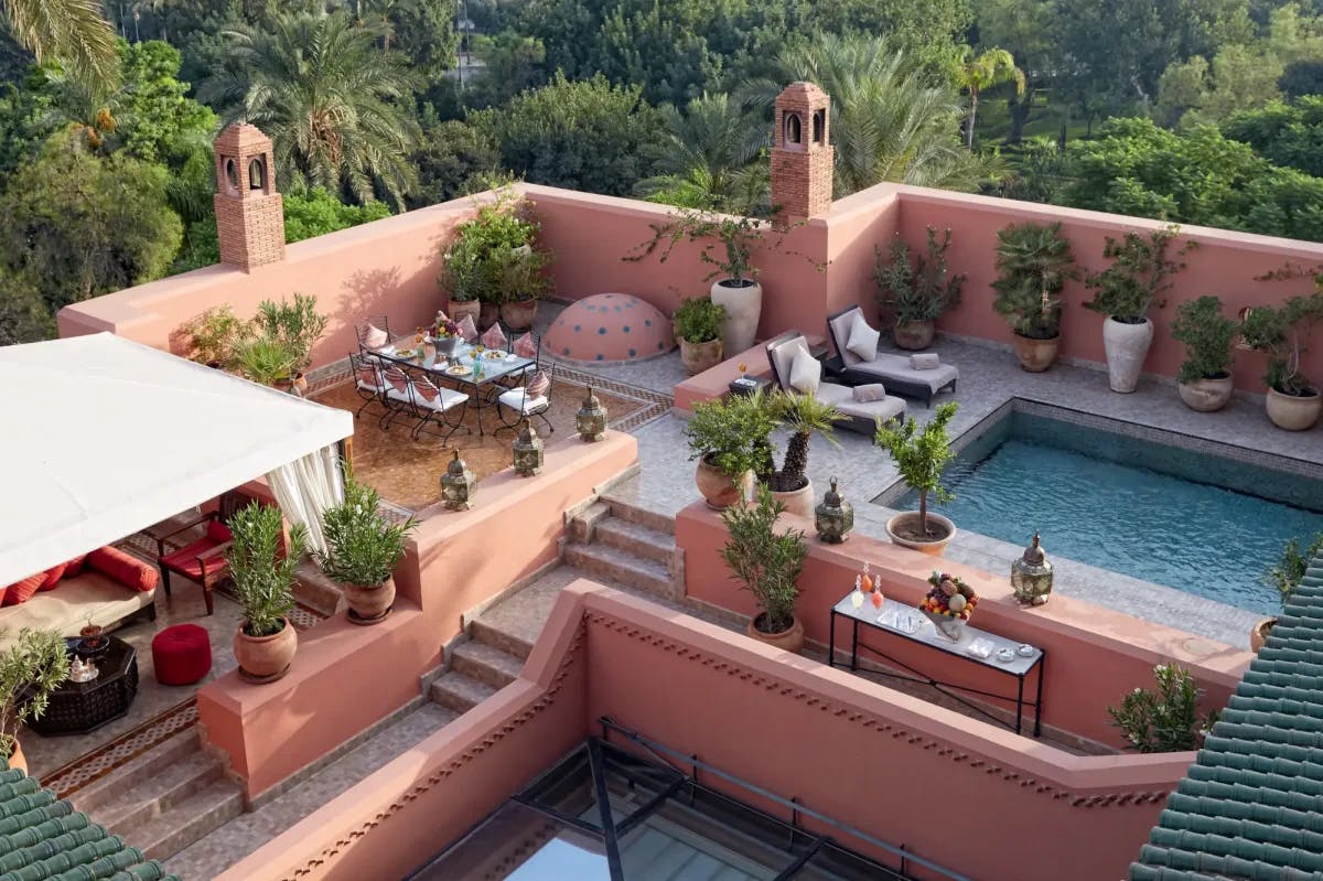 Within pink clay walls sit potted plants, a heated pool with lounge chairs, an ornate table setting and lounge area underneath a tent with drapes
