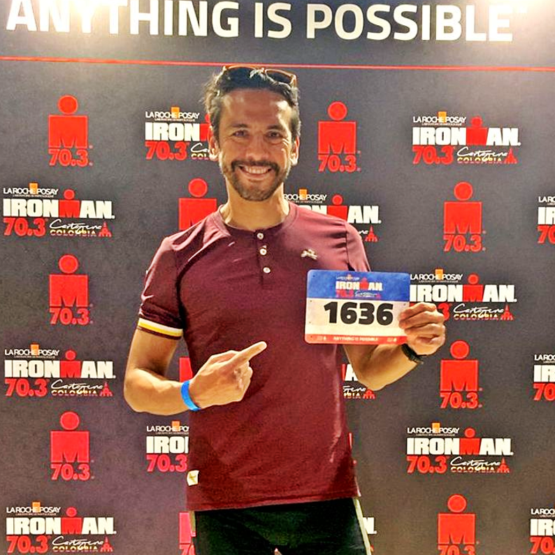 Gabriel Talavera in a red shirt holding a competitor's number with a background related to an Ironman