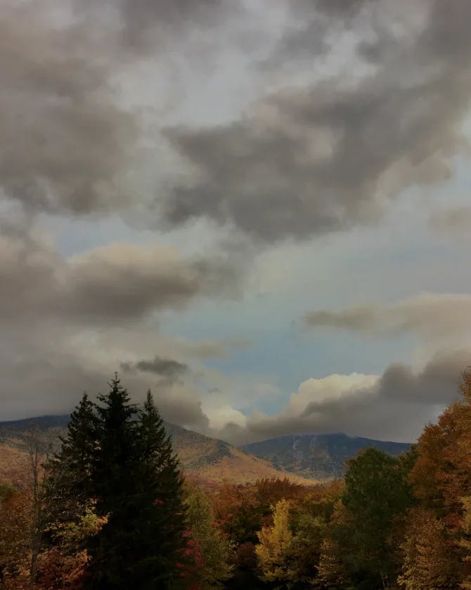 A view of a cloudy, moody sky with Autumnal trees and mountains in the bottom portion of the image. 