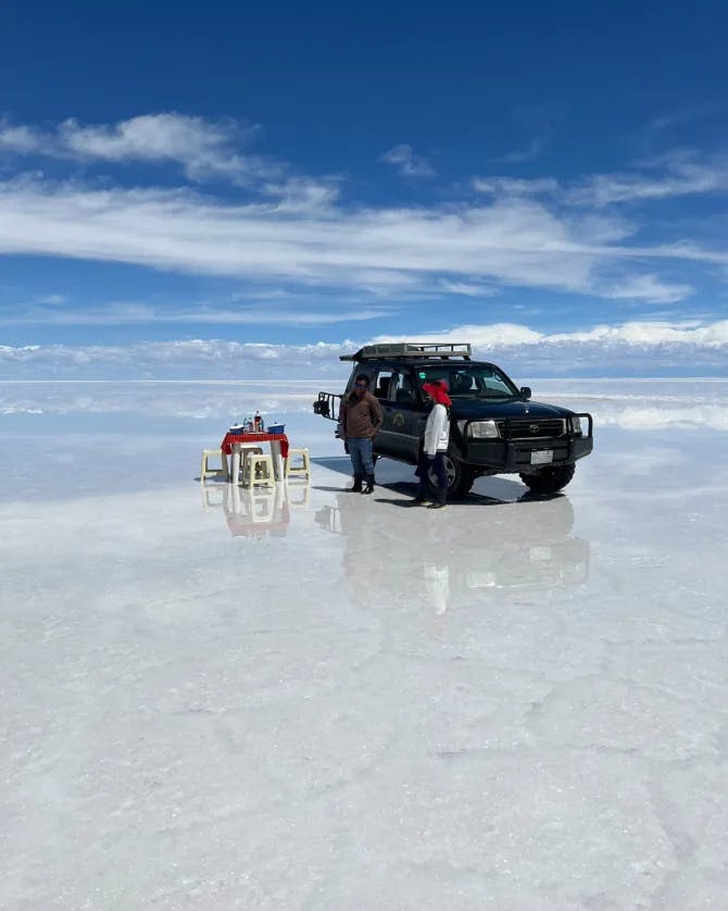 VIew of Uyuni Salt Flat with black jeep and a small table and chairs set up for a meal