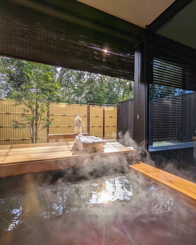 A view of a steam bath surrounded wooden architecture, towels and a fence surrounded by trees. 