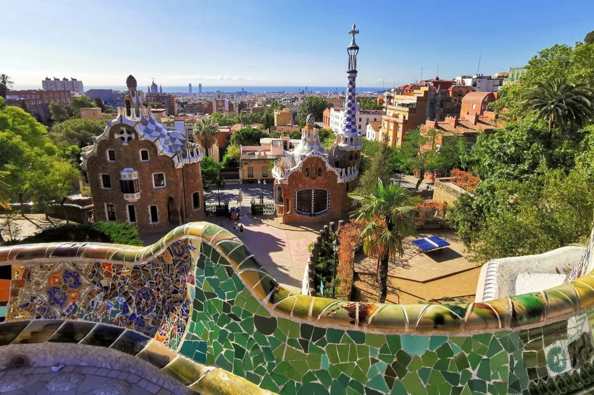 The style of Antoni Gaudí is evident in a prominently featured path wall. Behind, the historic skyline of Barcelona
