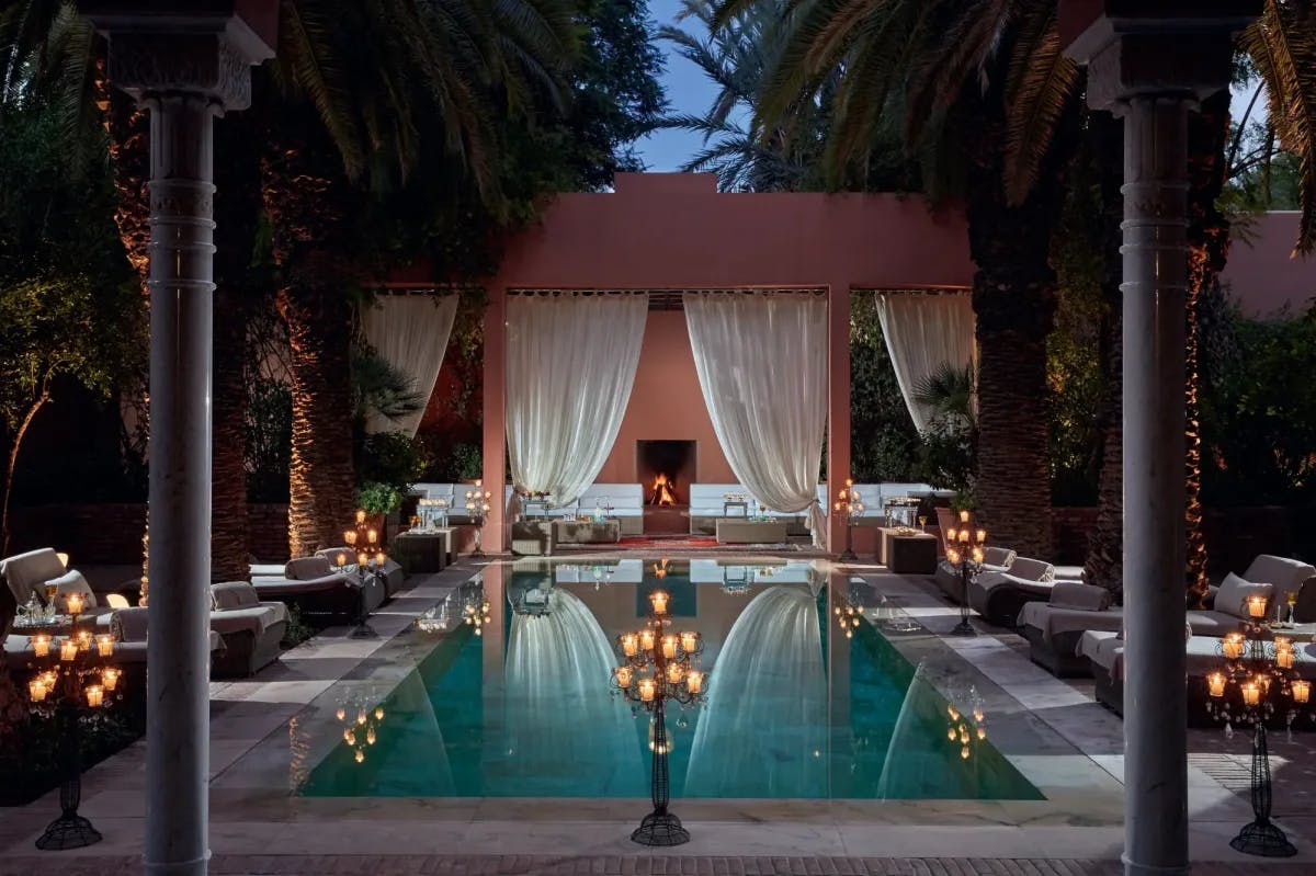 Lit by candles at twilight, lavish lounge seating surrounds a rectangular pool, with its surface reflecting white drapes attached to a private gazebo