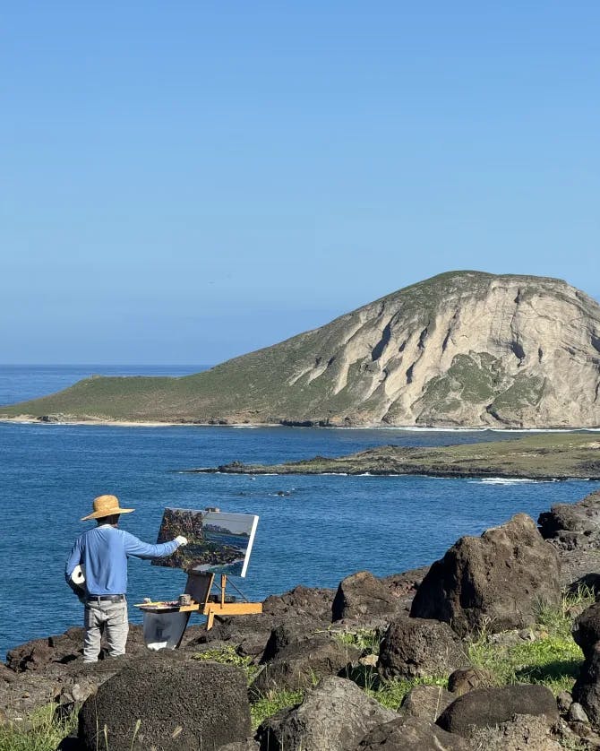 View of a man painting in with the ocean and mountain as the backdrop