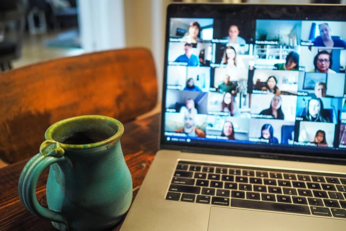 A laptop screen depicts dozens of people in a work conference next to a vintage coffee mug in a home office