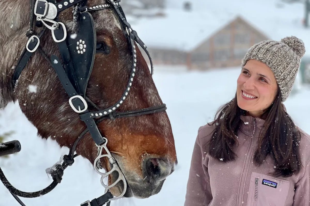 a woman in a white beanie stands next to a horse on a snowy day