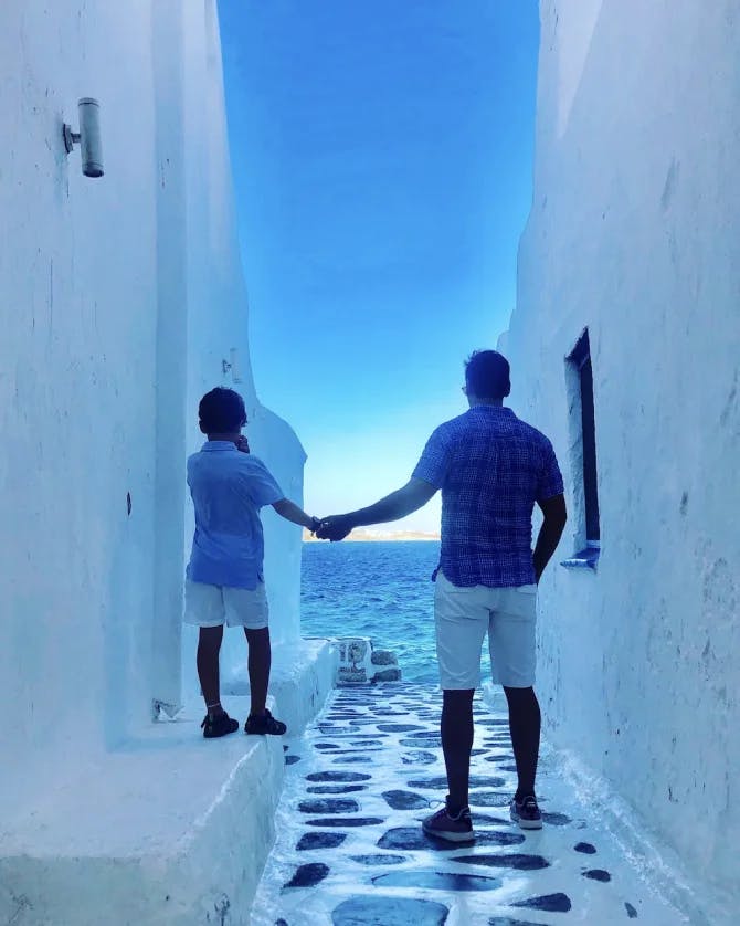 A man and a child standing in a Greek alley holding hands and looking out at the sea.