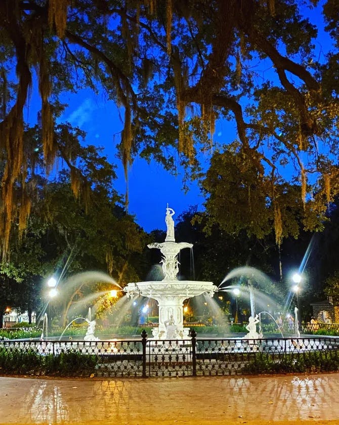 A beautiful photo at dusk of a lovely park with a large fountain and foliage in the distance.