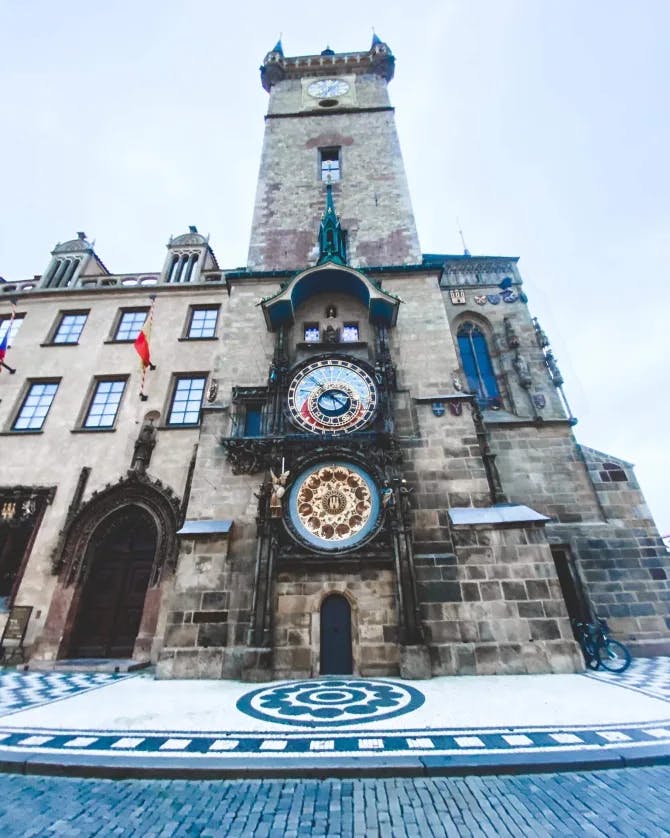 An old clock tower with stonework, windows, flags and a brick road. 