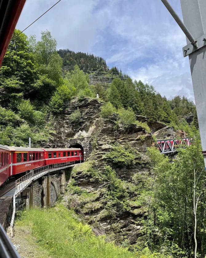 A red train on an elevated mountainside bridge traveling through a tunnel.
