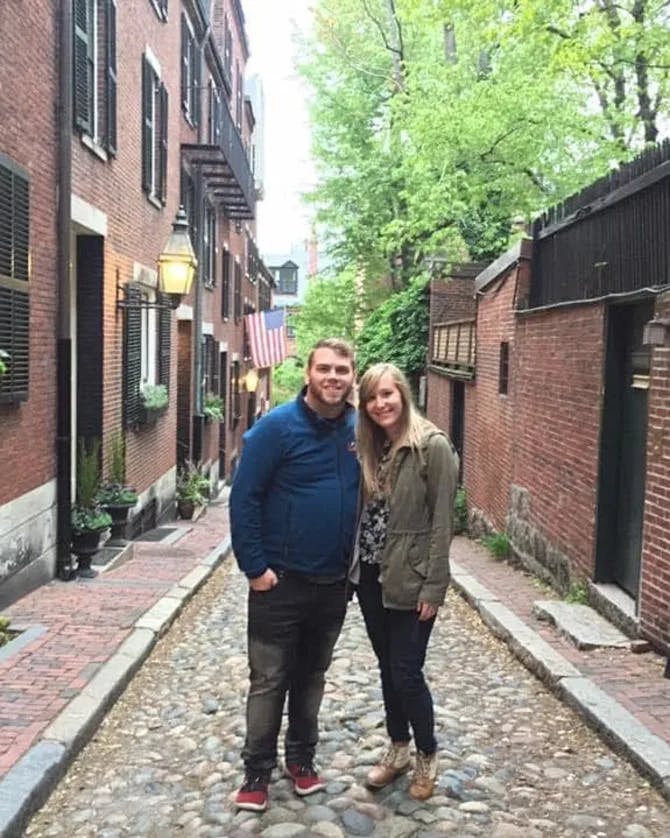 Elizabeth and a partner posing for a photo in a narrow brick filled alleyway. There are red-brick buildings on the left and ride sides, green planters and trees in the background. 
