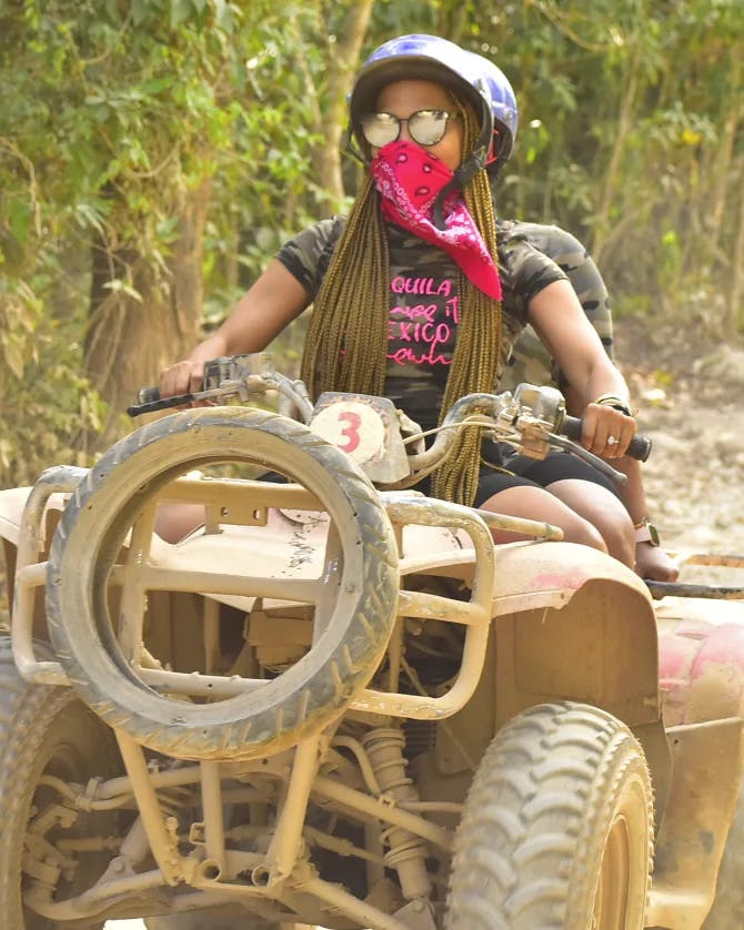 A person driving a dirt covered ATV outside. There are trees and brush in the background. 