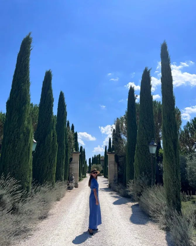 Travel advisor Sharon standing on a driveway in a blue dress and hat with tall trees both sides