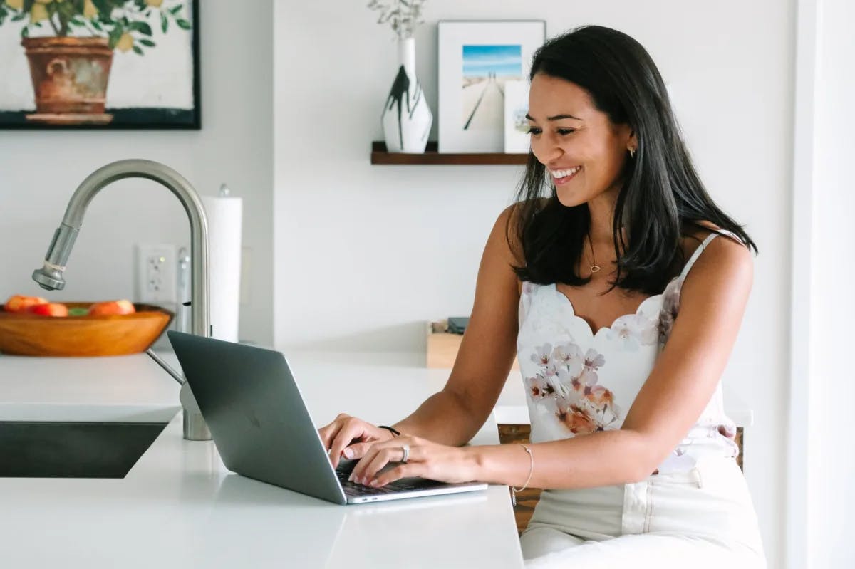 a woman in white pant and a white floral tank top works on her laptop at a kitchen counter