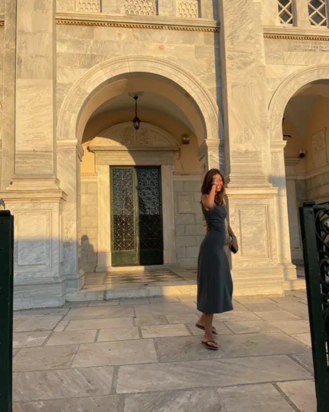 A woman wearing a black dress and posing in front of a beautiful old stone building with archways illuminated at sunset. 