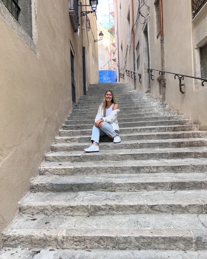 A woman wearing a white top and blue jeans on stone steps. There are stone walls and a metal railing to her right. 