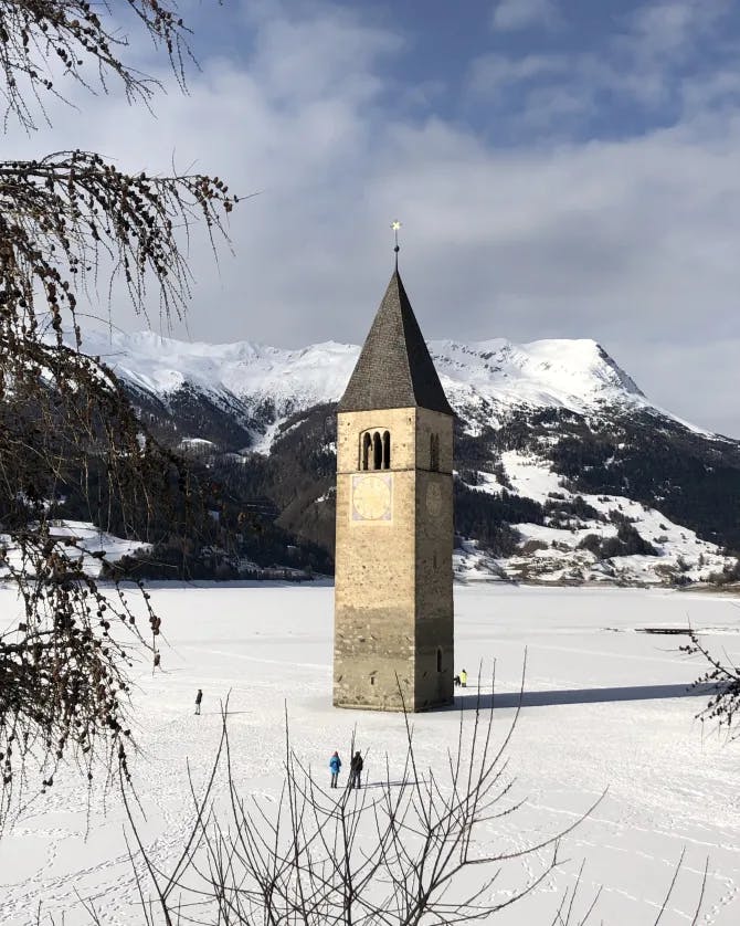 A stone tower in the middle of a snowy field with people walking, dried up trees and snowy mountains in the background. 