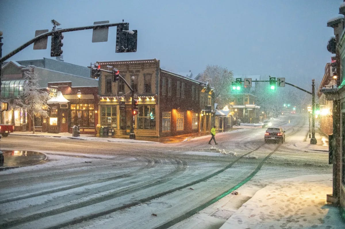 A view of a snowy main street with buildings, green street lights, cars, a pedestrian walking and exterior lights turned on. 