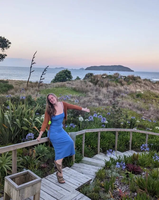A picture of Sonia wearing a blue dress and sitting on a wooden railing and staircase in front of a grassy meadow that leads to the sea