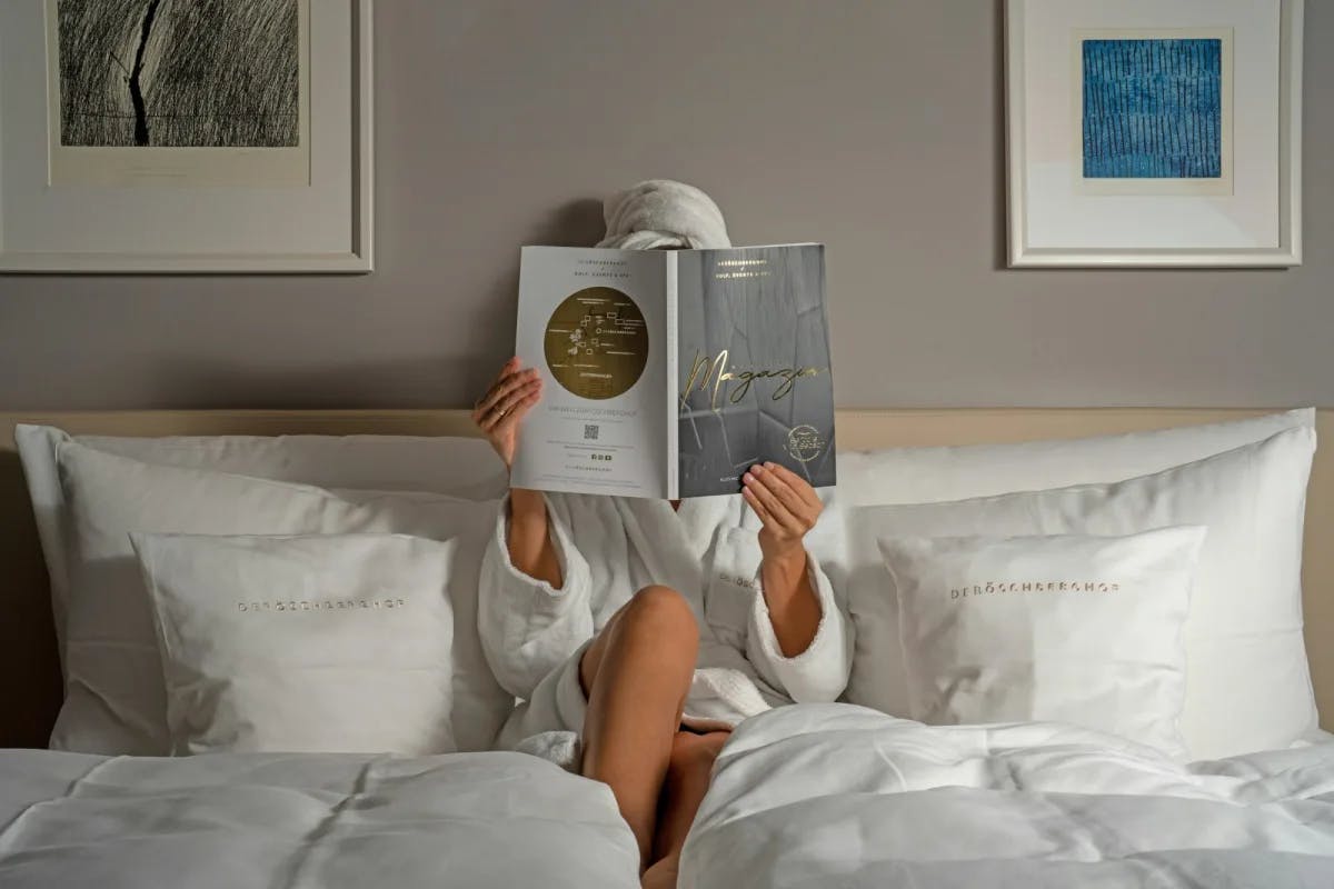 A woman in bathrobe sits on a plush, king-size hotel bed reading a luxe lifestyle magazine