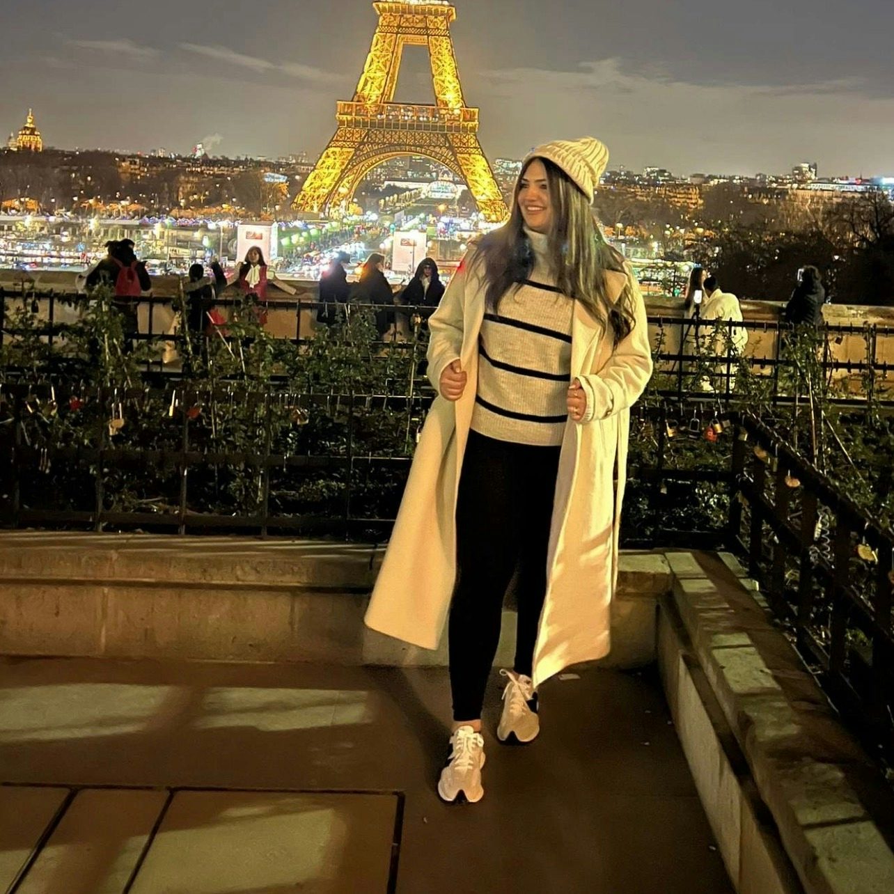 Riva Hanna in a white coat posing at night in front with the Eiffel Tower in view