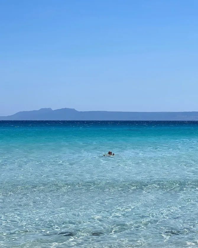 A beautiful view of crystal-clear sea water. There is a person swimming and a strip of land in the distance. 