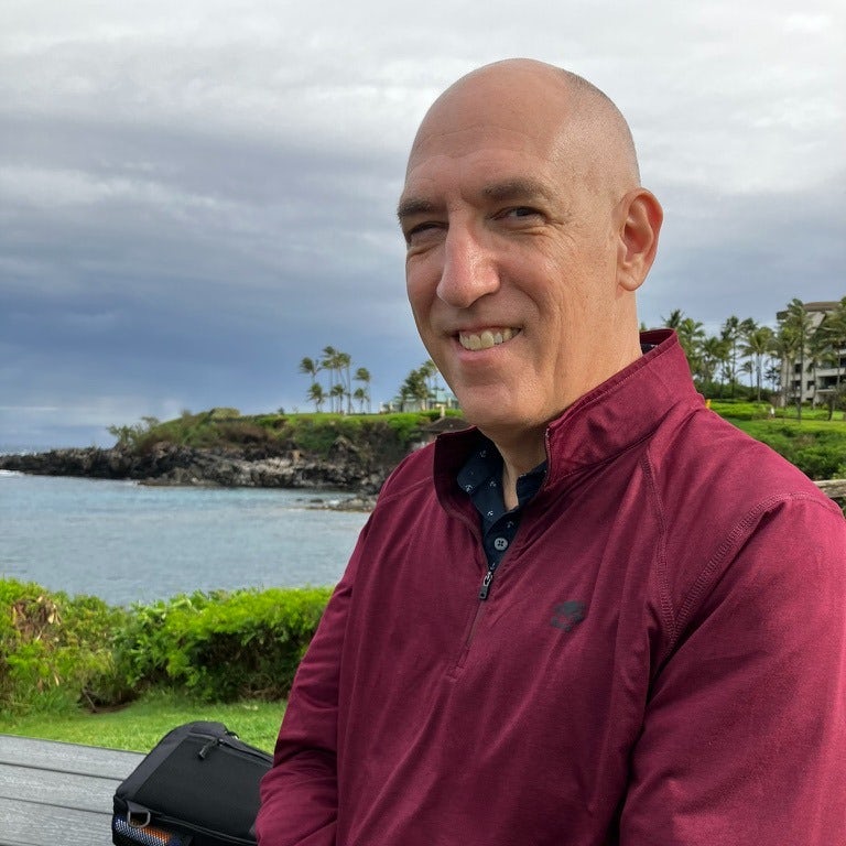 A man posing for a photo outside while wearing a maroon top. There is a body of water, palm trees and a green coastline in the background. 