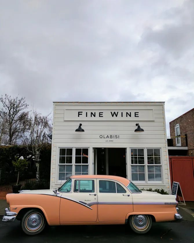 An old-fashioned orange and cream car parked in front of a white building with a sign that reads 'Fine Wine' in black lettering.