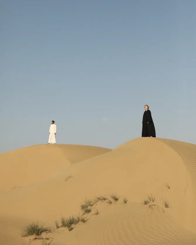 A view of two people, one wearing a white outfit and the other a black one, standing on top of a sand dune in the desert. 