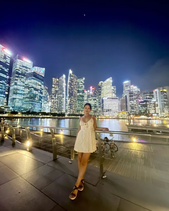 Cam posing in a white dress in front of a city skyline at nighttime