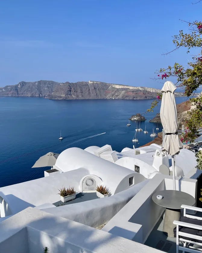 Santorini, with a small patio and chairs overlooking white cliffside houses and the ocean below, with an island in the distance.