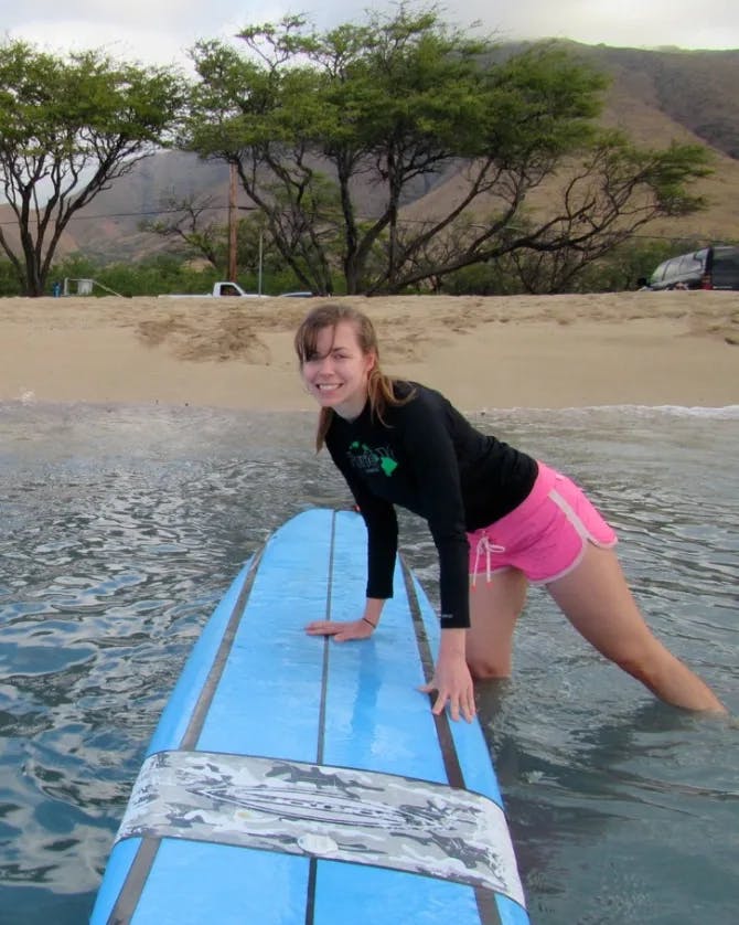 A woman posing with a blue surfboard in the shallow part of water with a beach, trees and mountains in the background. 