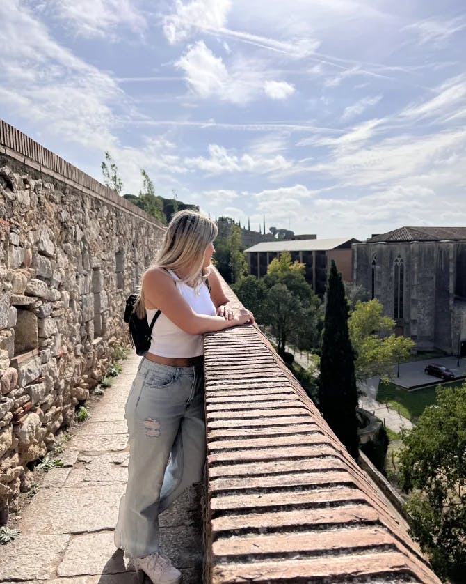 A woman wearing a white top and grey pants while leaning against a stone ledge. She is looking out towards a view of trees and a city scape. There is also a stone wall behind her.  