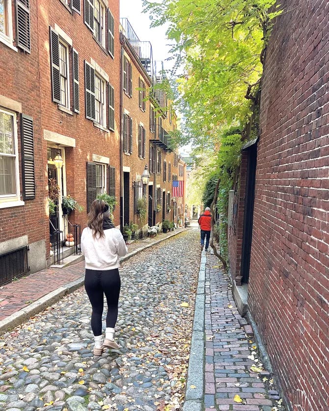 A brick road with two people walking down it. There are brick walls, black shutters, planters and trees in the surrounding areas.  
